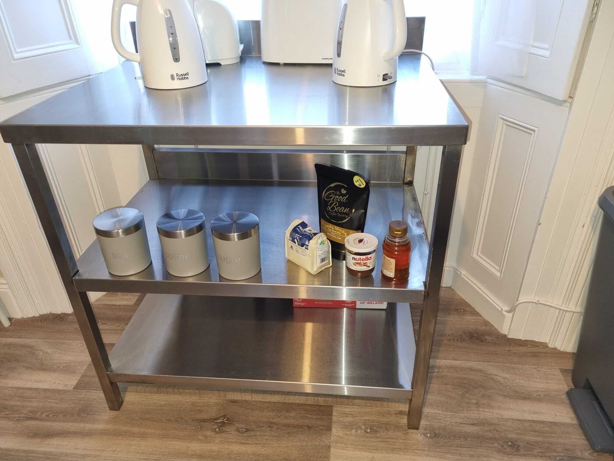 Eine Arbeitsplatte aus Edelstahl mit zwei Wasserkochern, drei Behältern mit der Aufschrift Tee, Kaffee und Zucker, einer Kaffeebohnentasche, einem Nutella-Glas und einem Honigglas.