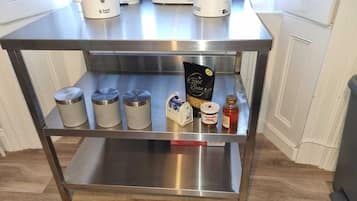 Eine Arbeitsplatte aus Edelstahl mit zwei Wasserkochern, drei Behältern mit der Aufschrift Tee, Kaffee und Zucker, einer Kaffeebohnentasche, einem Nutella-Glas und einem Honigglas.
