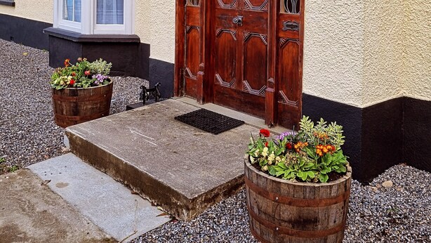 A wooden front door with a decorative glass panel, a small window with a white frame, and two wooden planters with flowers. - The Monkey Puzzle (Bunclody)