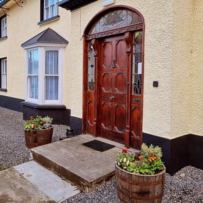 A wooden front door with a decorative glass panel, a small window with a white frame, and two wooden planters with flowers. - The Monkey Puzzle (Bunclody)