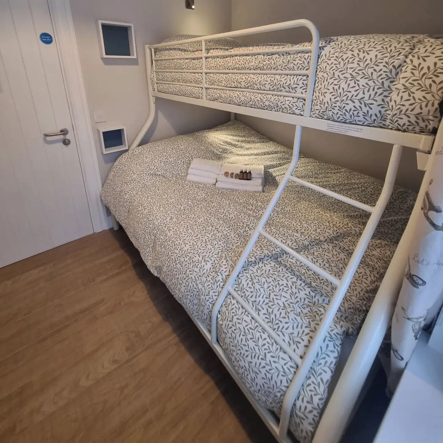 Una cama individual con ropa de cama de diseño de hojas, una mesita de noche con una botella y una caja, y un pequeño estante.