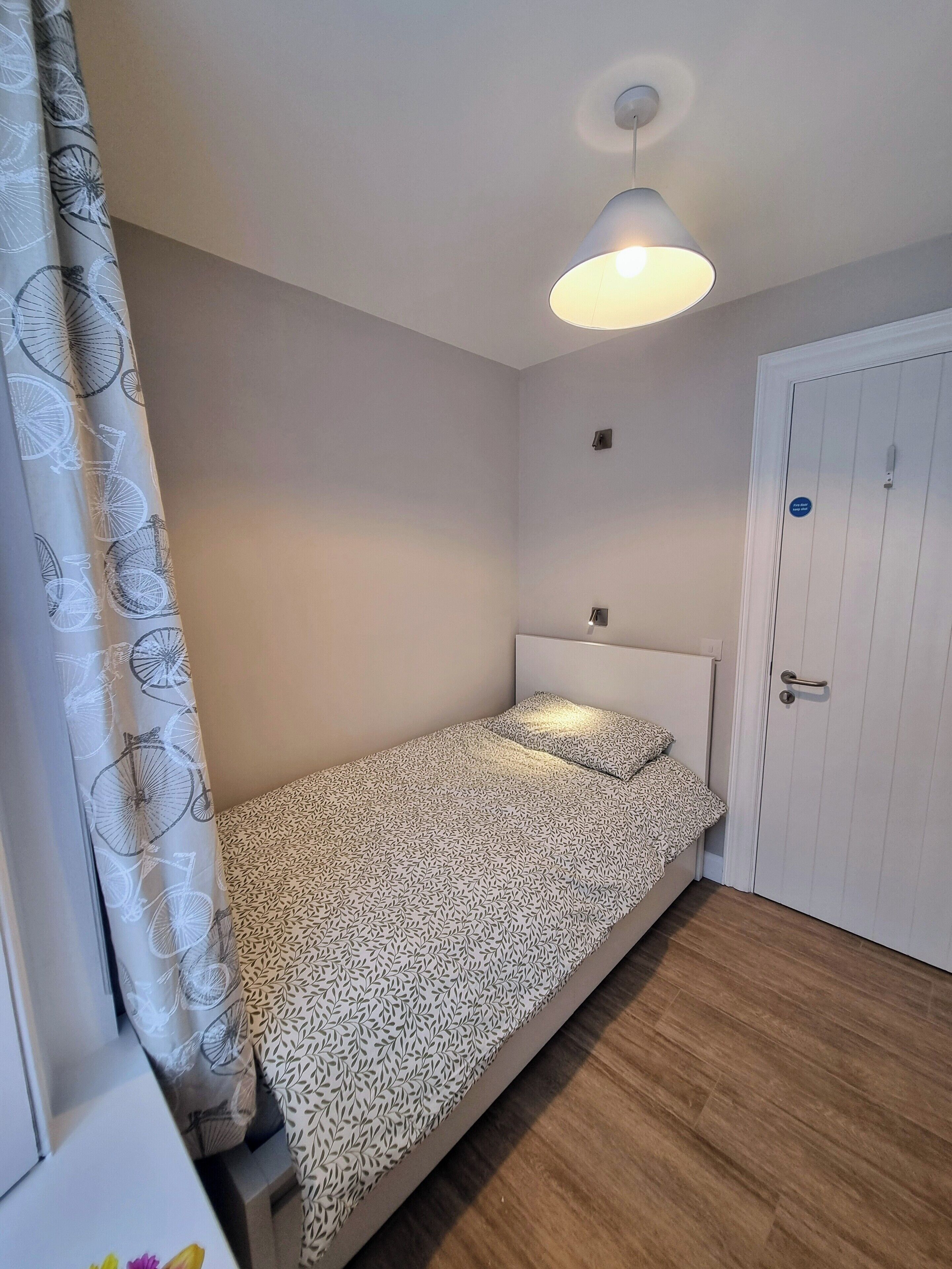 Una cama individual con una colcha estampada en una habitación con suelo de madera, una puerta blanca y una lámpara en el techo.