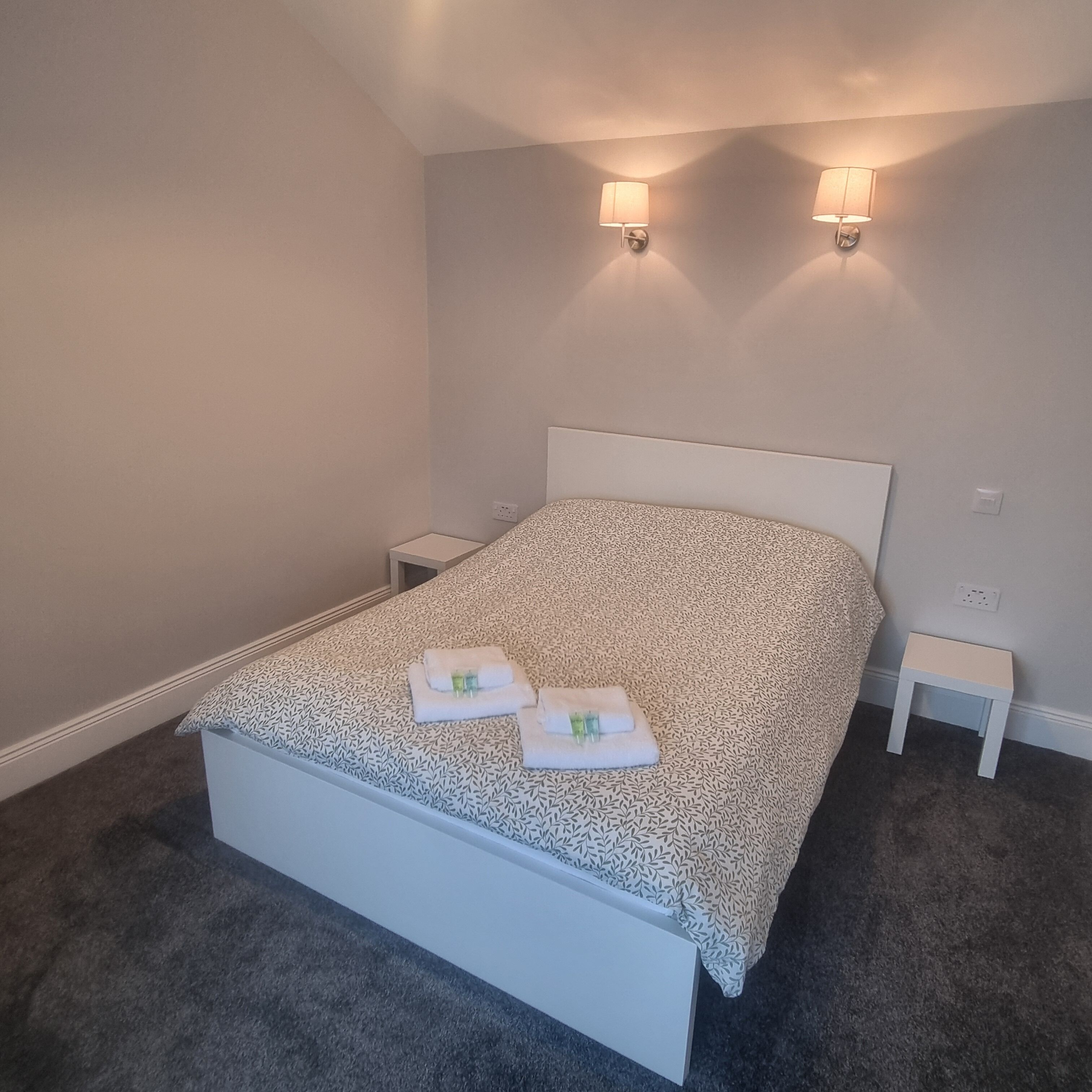 A neatly made bed with a patterned bedspread, two bedside tables, and wall-mounted lights.