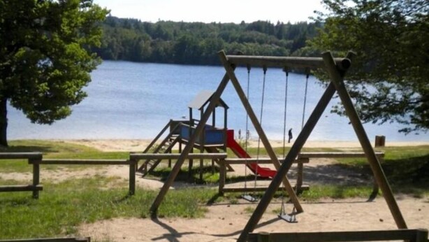 Et legesæt i træ med gyngestativ og rutchebane ved en sø med sandstrand og træer i baggrunden.