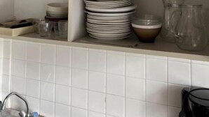 Et køkken med hvid flisebeklædt væg, hvide hylder med stablet service, en kaffemaskine og en vask.