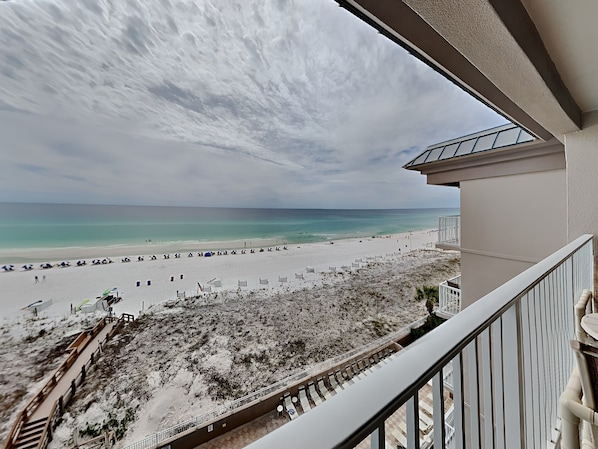 Enjoy a serene beachfront view from this balcony, overlooking a sandy beach with umbrellas and beach chairs.