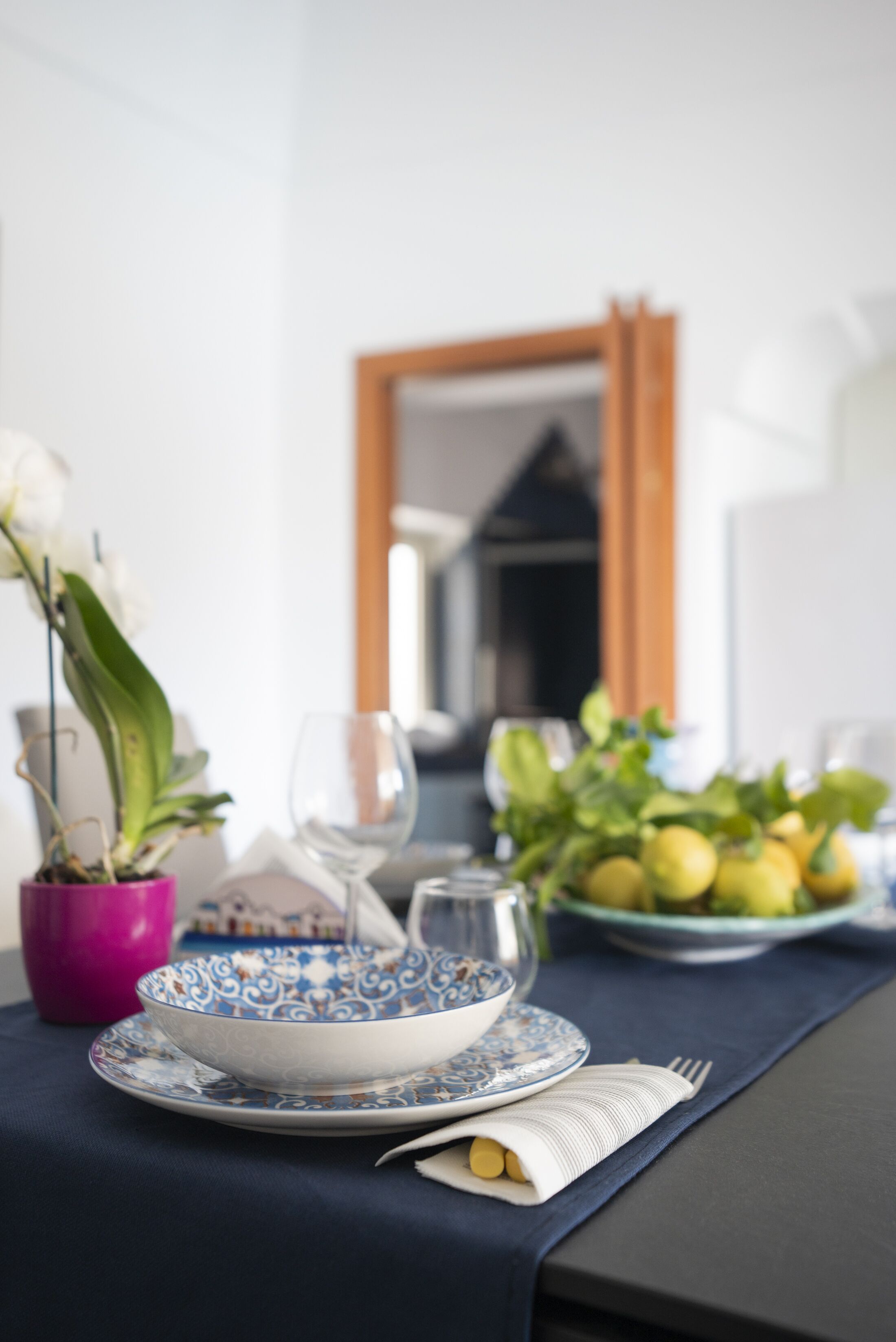 A dining table set with a blue and white patterned bowl, a pink vase with white flowers, and a plate of lemons.