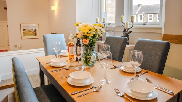 Een eettafel is gedekt met witte borden, bestek en wijnglazen, omringd door grijze fauteuils. Op de tafel staat een vaas met gele bloemen en kaarsen.