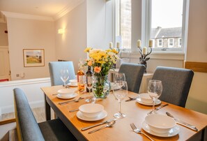 Een eettafel is gedekt met witte borden, bestek en wijnglazen, omringd door grijze fauteuils. Op de tafel staat een vaas met gele bloemen en kaarsen.