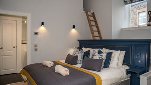 Een opgemaakt bed met een blauw hoofdbord, twee wandlampen, een houten ladder en een raam met uitzicht op een stenen gebouw.