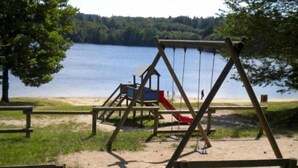 A wooden swing set and slide near a lake with a forested background. - Gite 3 Personnes (Ambrugeat)