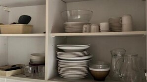 A kitchen with white tiled walls, a sink, and shelves holding dishes, bowls, and a pitcher. - Gite 3 Personnes (Ambrugeat)