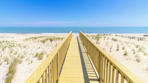 Ein hölzerner Steg führt zu einem Strand mit Dünen und blauem Himmel.
