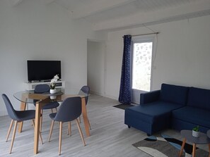 Een moderne woonkamer met een blauwe bank, een glazen eettafel met stoelen, een flatscreen-televisie en een groot raam met decoratieve gordijnen.