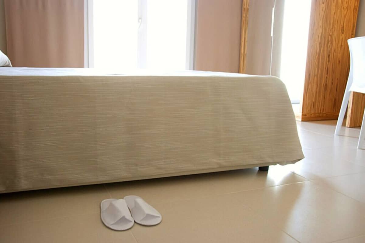 Een opgemaakt bed met een witte dekbedovertrek en een paar witte pantoffels op de vloer.