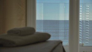 A room with a bed, a window with a view of the sea, and a balcony with a railing.
