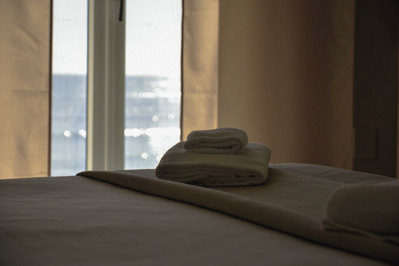 A neatly made bed with a folded towel on top, a window with curtains, and a view of a cityscape.