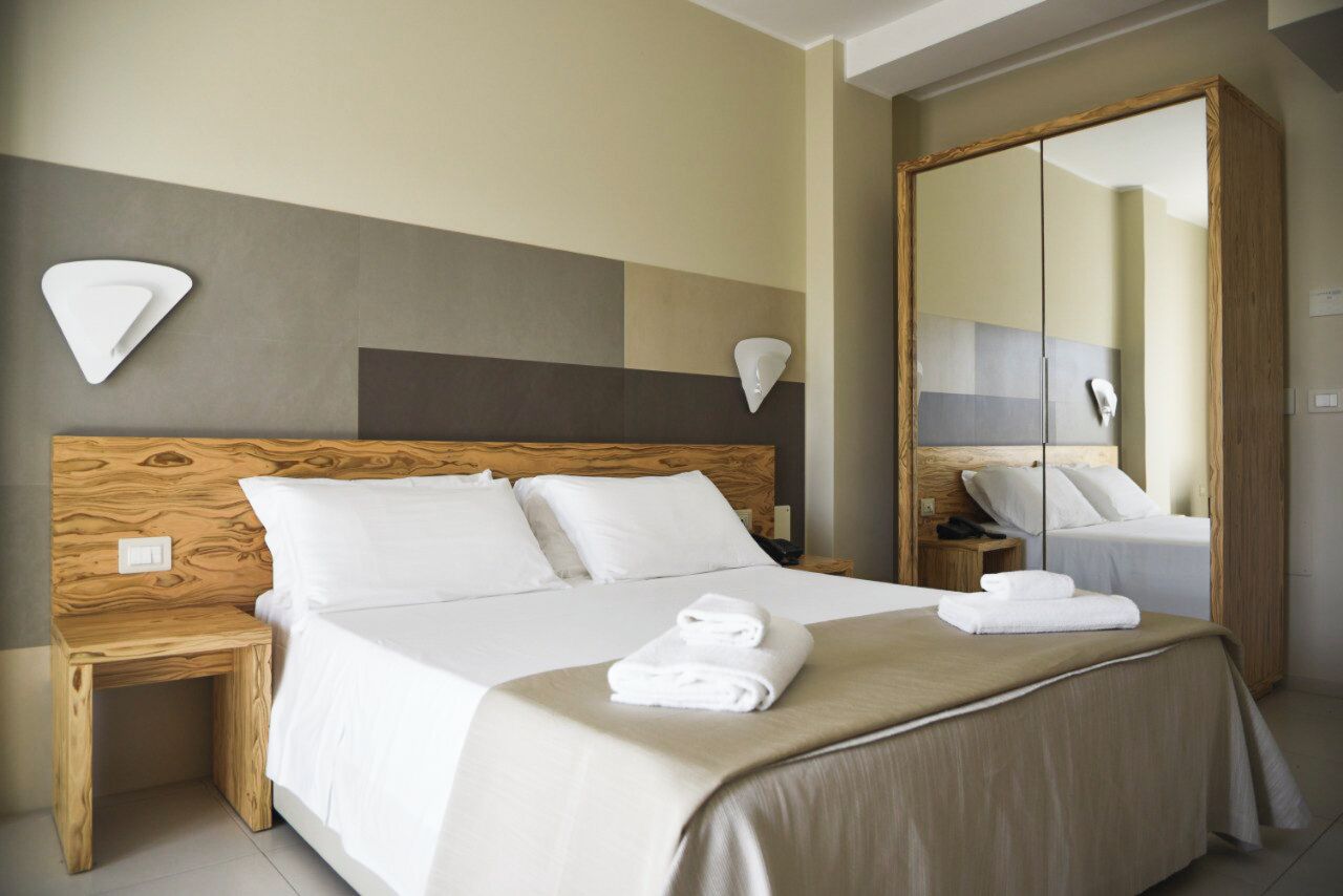 Une chambre d’hôtel avec une tête de lit en bois, un grand miroir et un lit bien fait, avec du linge de lit et des serviettes blanches.