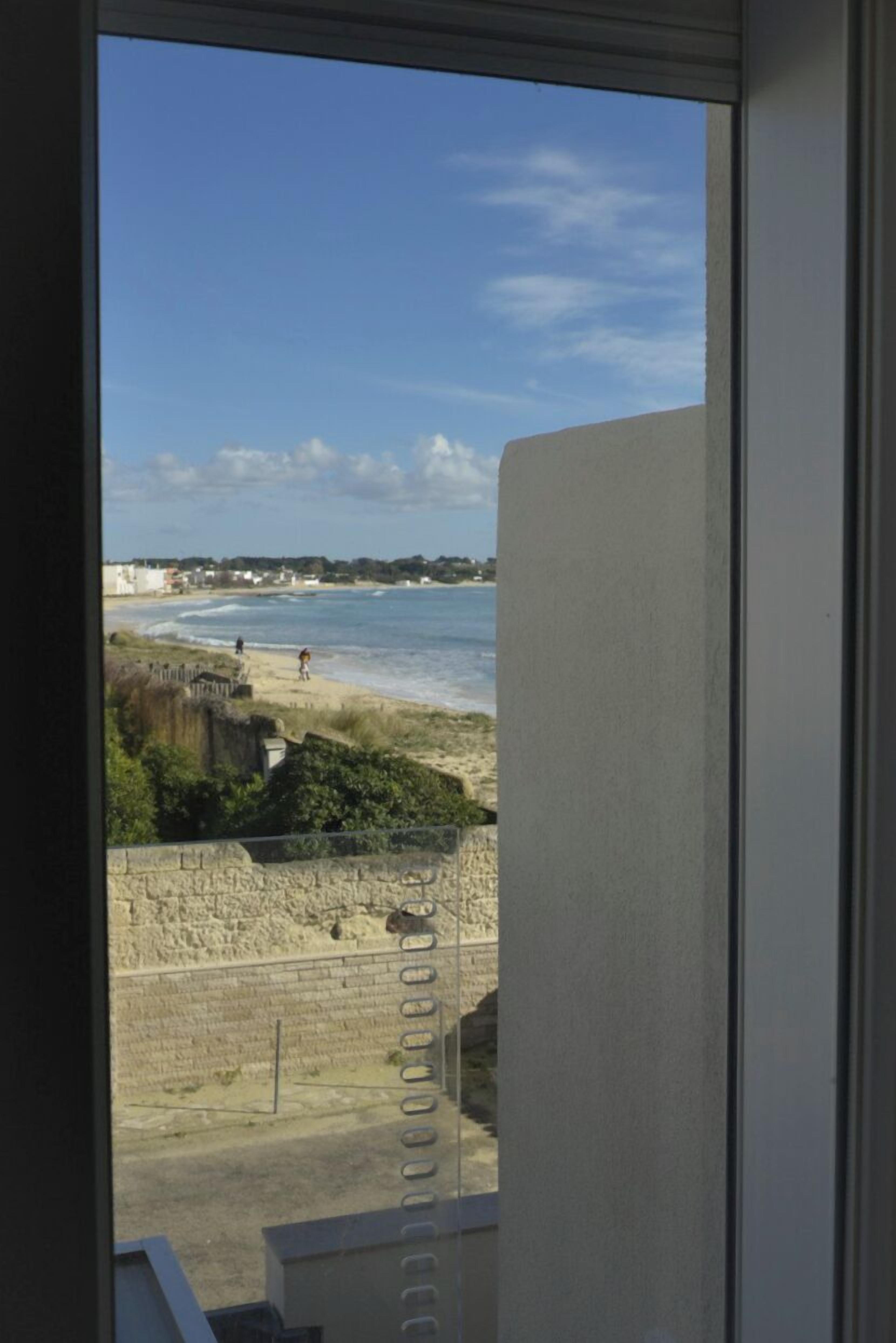 Vista dalla finestra su una spiaggia, con un muro di pietra e edifici sullo sfondo.