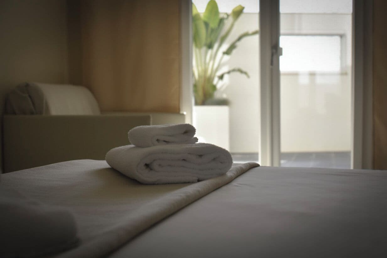 A neatly made bed with a folded towel on top, a bedside table, and a view of a balcony with a plant.
