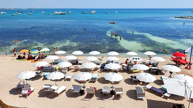 A beach with rows of sunbeds and umbrellas, boats in the water, and clear blue skies.