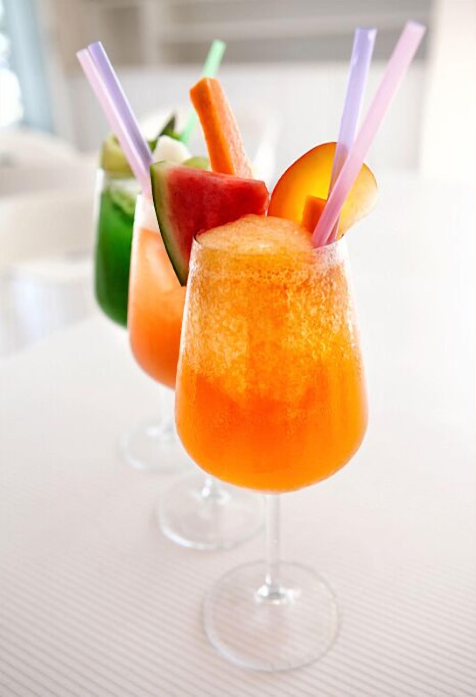 Dos cócteles con adornos de frutas y pajitas en vasos transparentes sobre una superficie blanca.