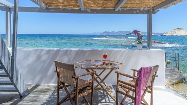 A covered outdoor seating area with a table and chairs, overlooking a coastal view with clear blue water and distant landforms. - Esperanza Boathouse (Mandrakia)