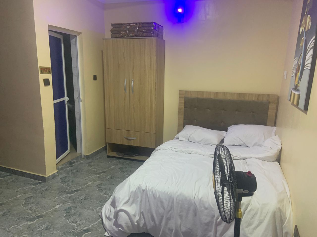 A bedroom with a bed, a wooden wardrobe, a fan, and a door with a sign.