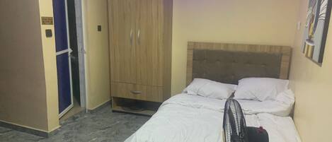 Une chambre avec un lit, une armoire en bois, un ventilateur et une porte sur laquelle est apposée une pancarte.