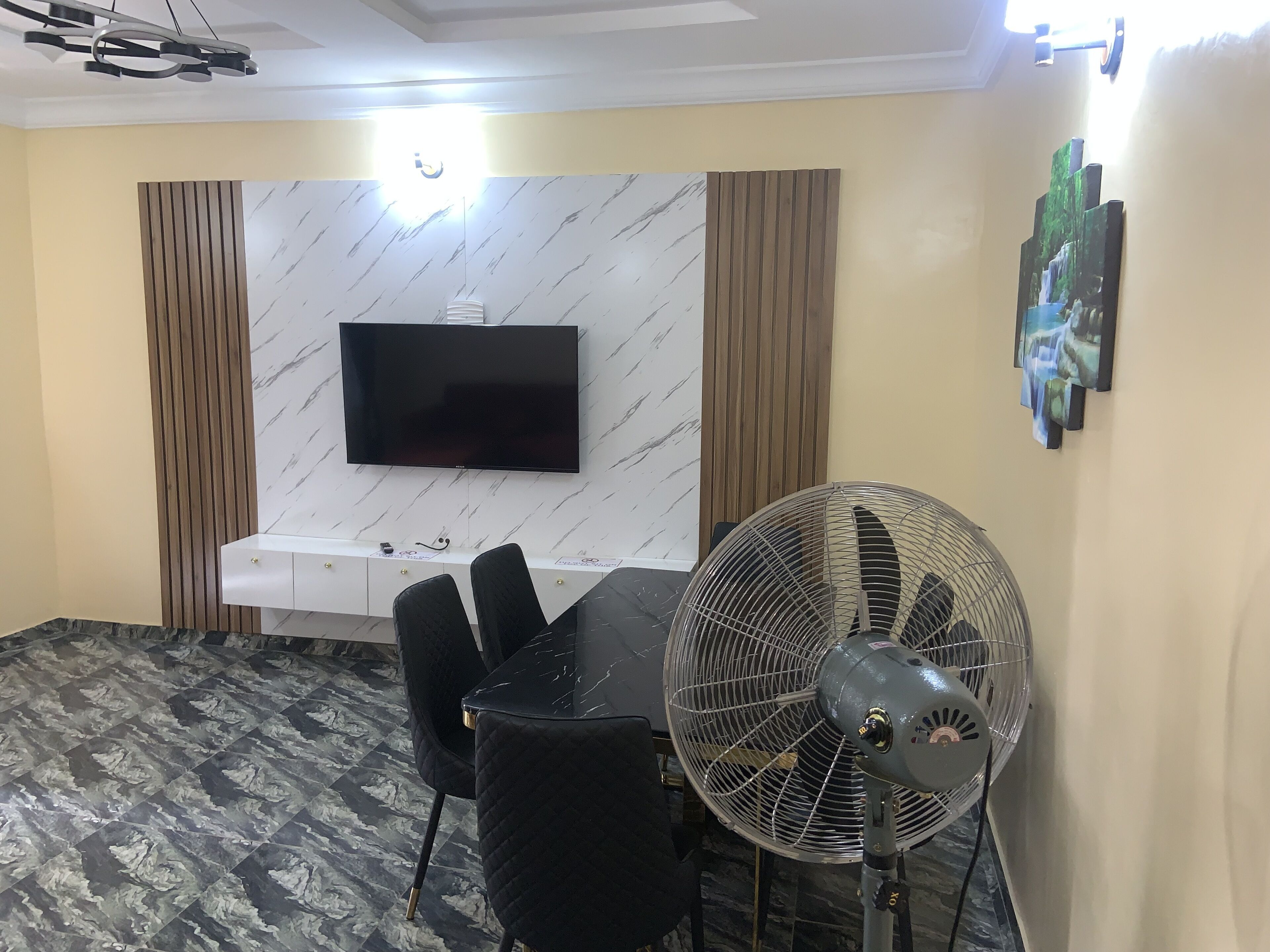 Un espace repas moderne avec un téléviseur à écran plat fixé à un mur en marbre, une table à manger avec des chaises noires, un ventilateur sur pied et un élément décoratif mural.