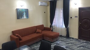 A room with a brown sectional sofa, a glass-top dining table with four chairs, a ceiling light fixture, an air conditioning unit, and a door with a security lock. - Blue Icon Apartment  (Loburo)