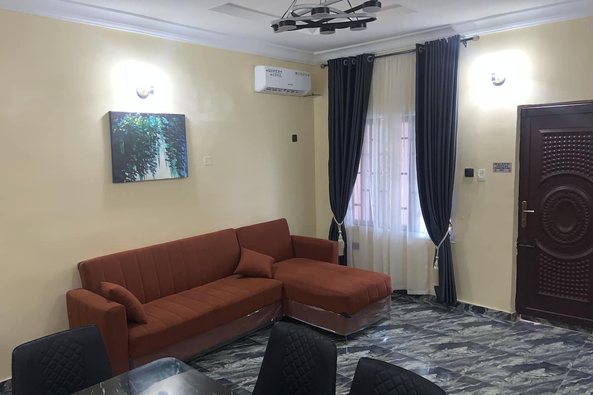 A room with a brown sectional sofa, a glass-top dining table with four chairs, a ceiling light fixture, an air conditioning unit, and a door with a security lock.