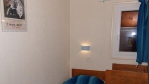 Un petit logement composé d’une seule pièce, comprenant deux lits en bois, une fenêtre avec des rideaux, un luminaire fixé au mur et une affiche accrochée au mur.