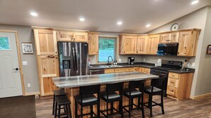 A modern kitchen with wooden cabinets, a stainless steel refrigerator, a large island with bar stools, and a built-in oven. - Lake Nokomis cabin (Tomahawk)