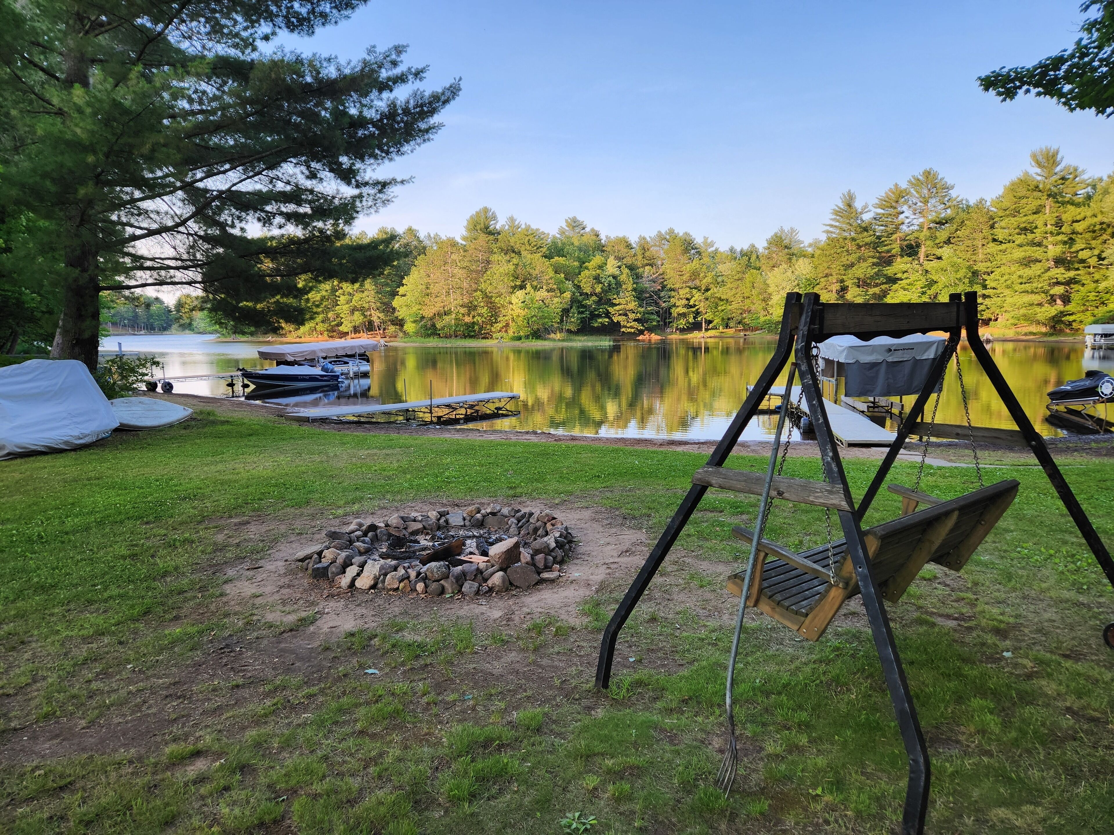 Un espace de camping au bord du lac, équipé d’un foyer, d’une balançoire et de bateaux amarrés près de l’eau.