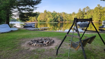 Un espace de camping au bord du lac, équipé d’un foyer, d’une balançoire et de bateaux amarrés près de l’eau.