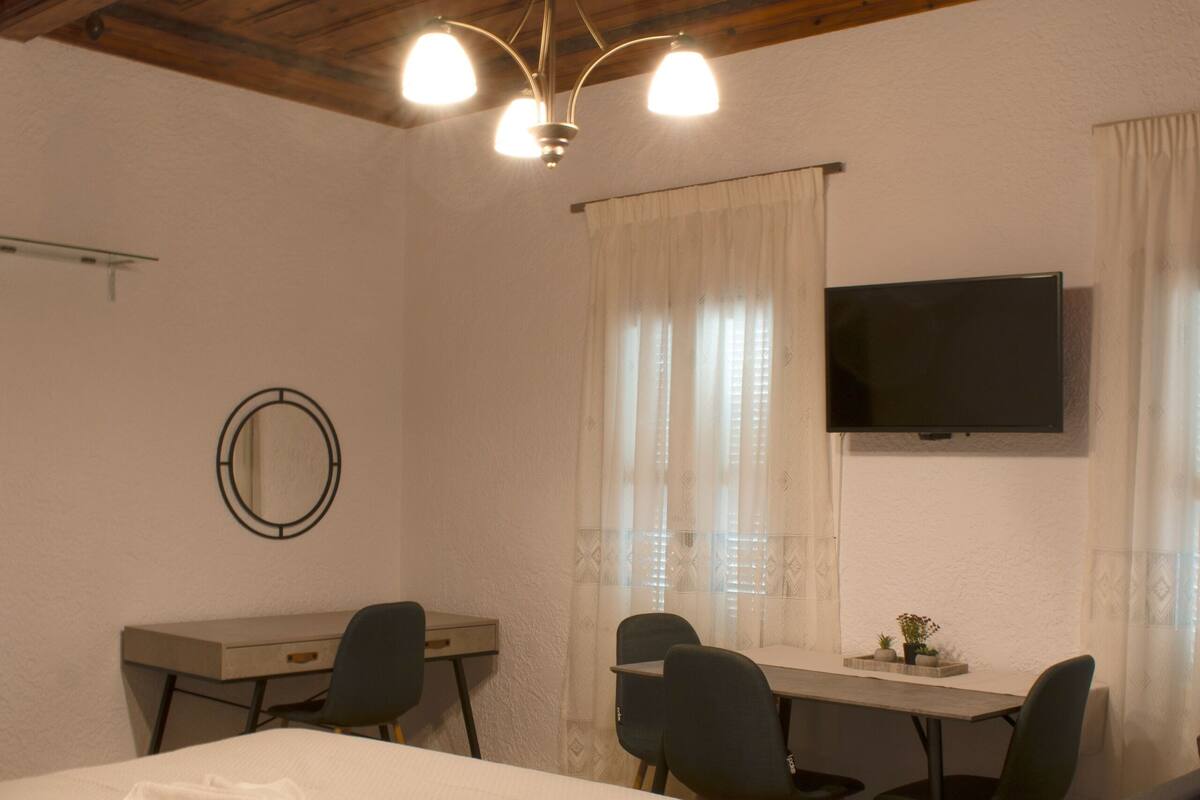 A hotel room with a bed, a desk, two chairs, a TV, and a ceiling light fixture.