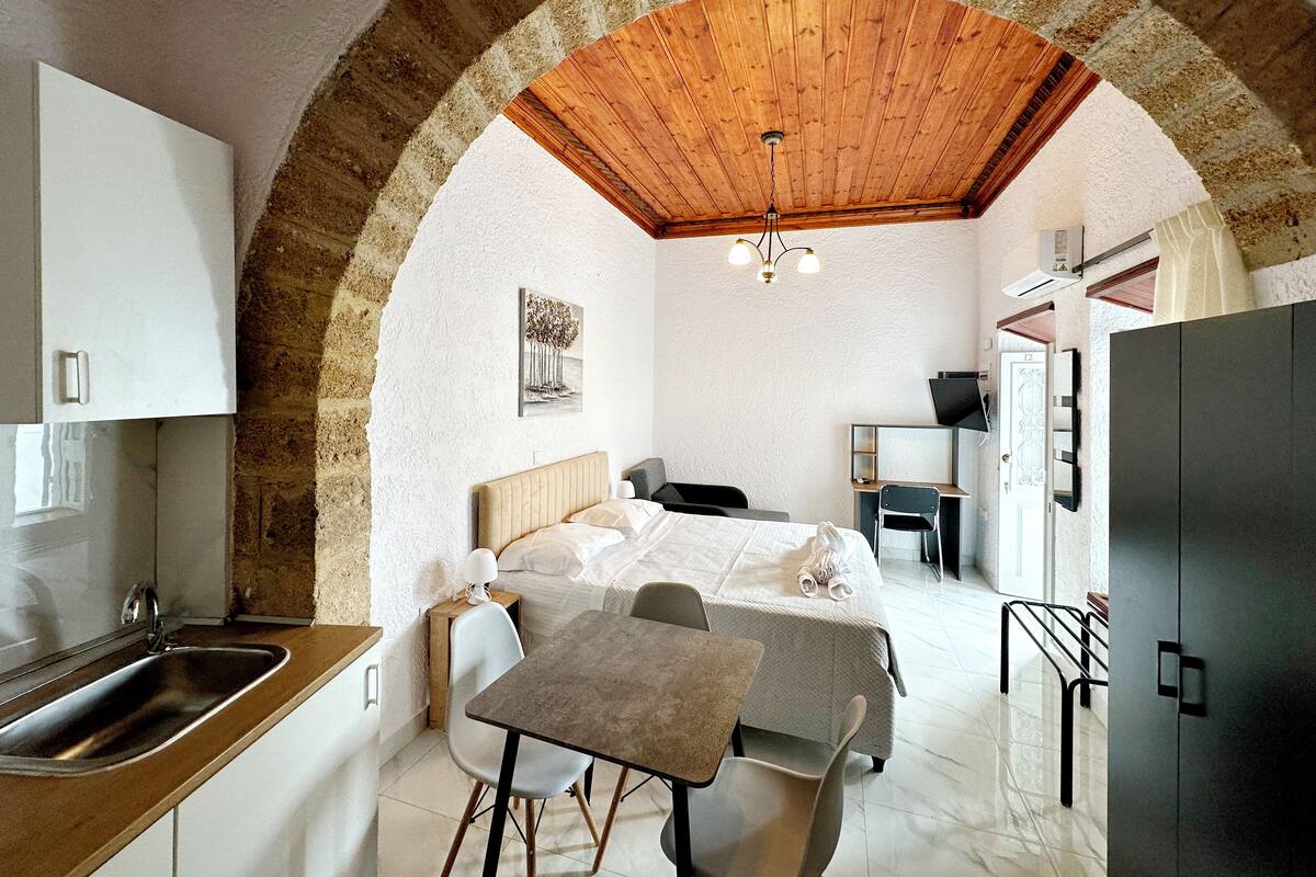A compact hotel room with a kitchen area, a bed with a headboard, a small table, and a chair.