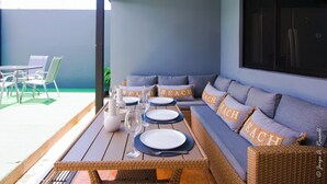Un'area pranzo all'aperto coperta con un tavolo apparecchiato per due, un divano in vimini con cuscini con la scritta 'BEACH' e vista sul patio.