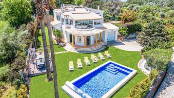 Una amplia casa de varios pisos con piscina, rodeada de un césped cuidado y palmeras.