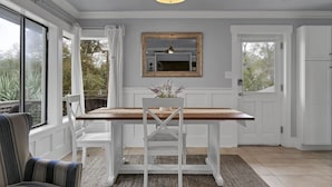 A dining area with a wooden table, white chairs, a mirror, and a view through large windows.