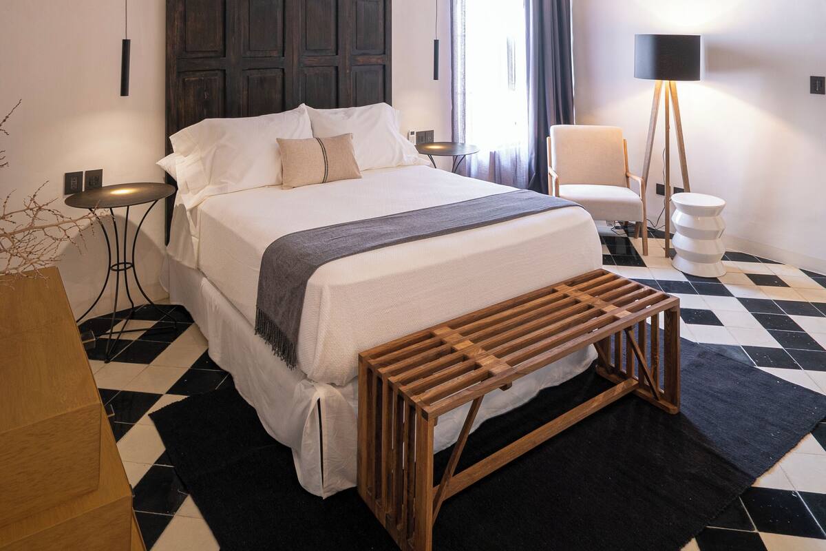 A modern bedroom with a large bed, a bedside table, a lamp, and a bench.