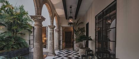 A corridor with a checkered floor, columns, and potted plants.