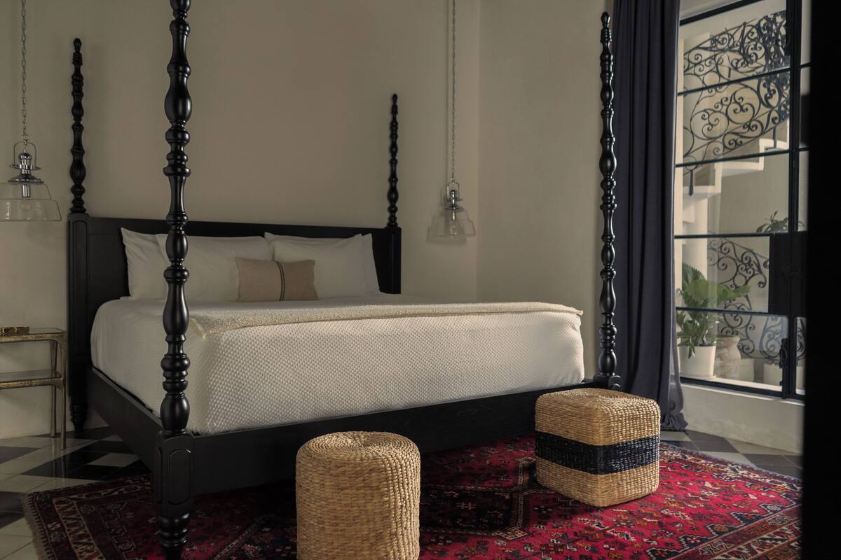 A four-poster bed with a dark wooden frame, a white bedspread, and two woven ottomans on a red patterned rug.