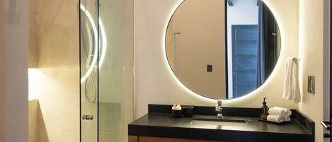 Une salle de bain avec une douche à parois de verre, un miroir rond avec un éclairage intégré, un meuble-lavabo en bois et un carrelage à motifs carrés.