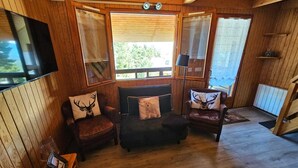 Een gezellig houten interieur met een groot raam met zonwering, een flatscreen televisie aan de muur en twee leren fauteuils met kussens met hertenmotief.