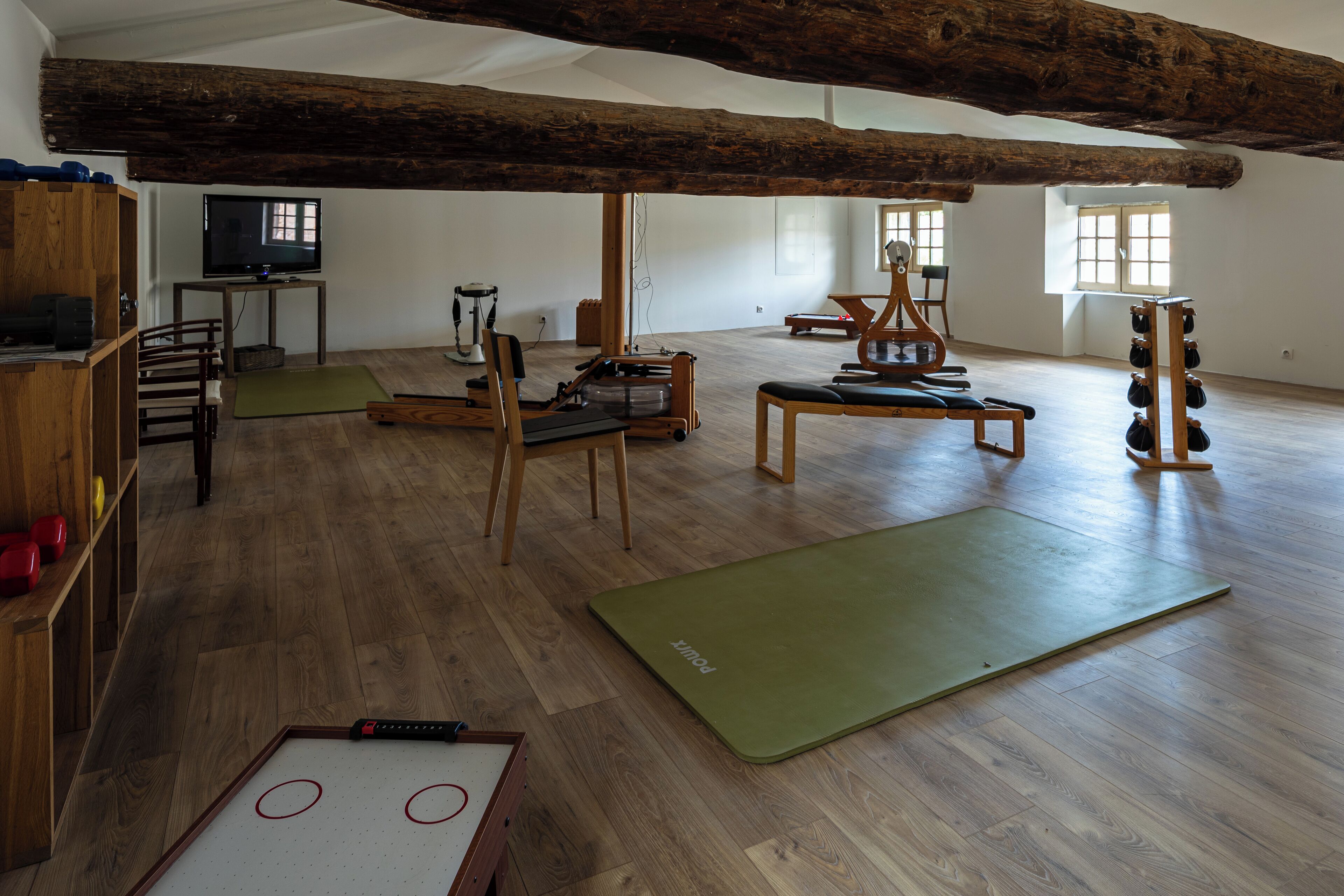 A spacious room with wooden floors, a large wooden beam ceiling, a treadmill, exercise mats, and a television.