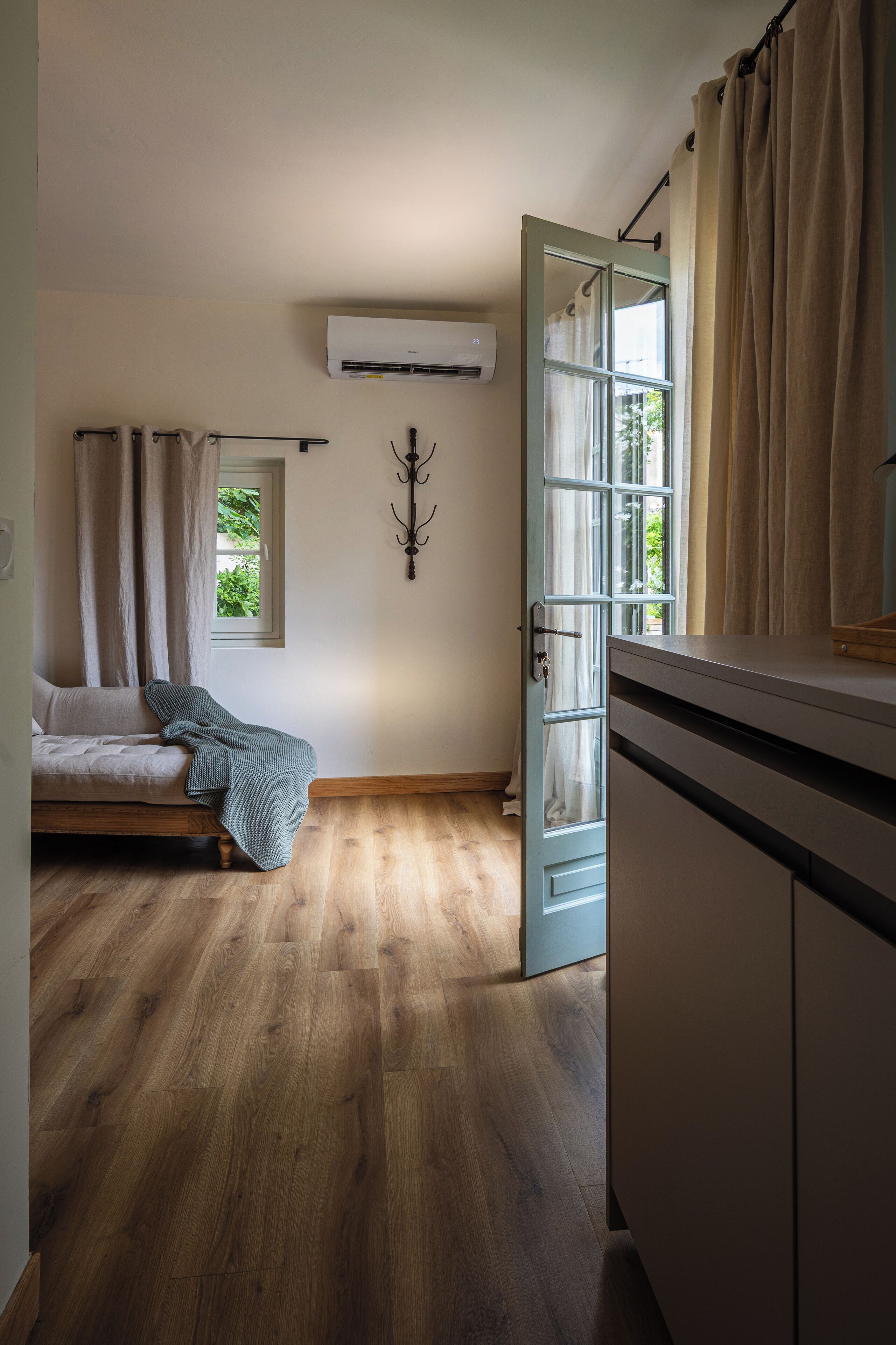 A room with a bed, a window with curtains, an air conditioning unit, and a wooden floor.