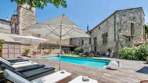 Un área junto a la piscina con sillones de descanso y sombrillas, un edificio de piedra y una piscina azul cristalina.
