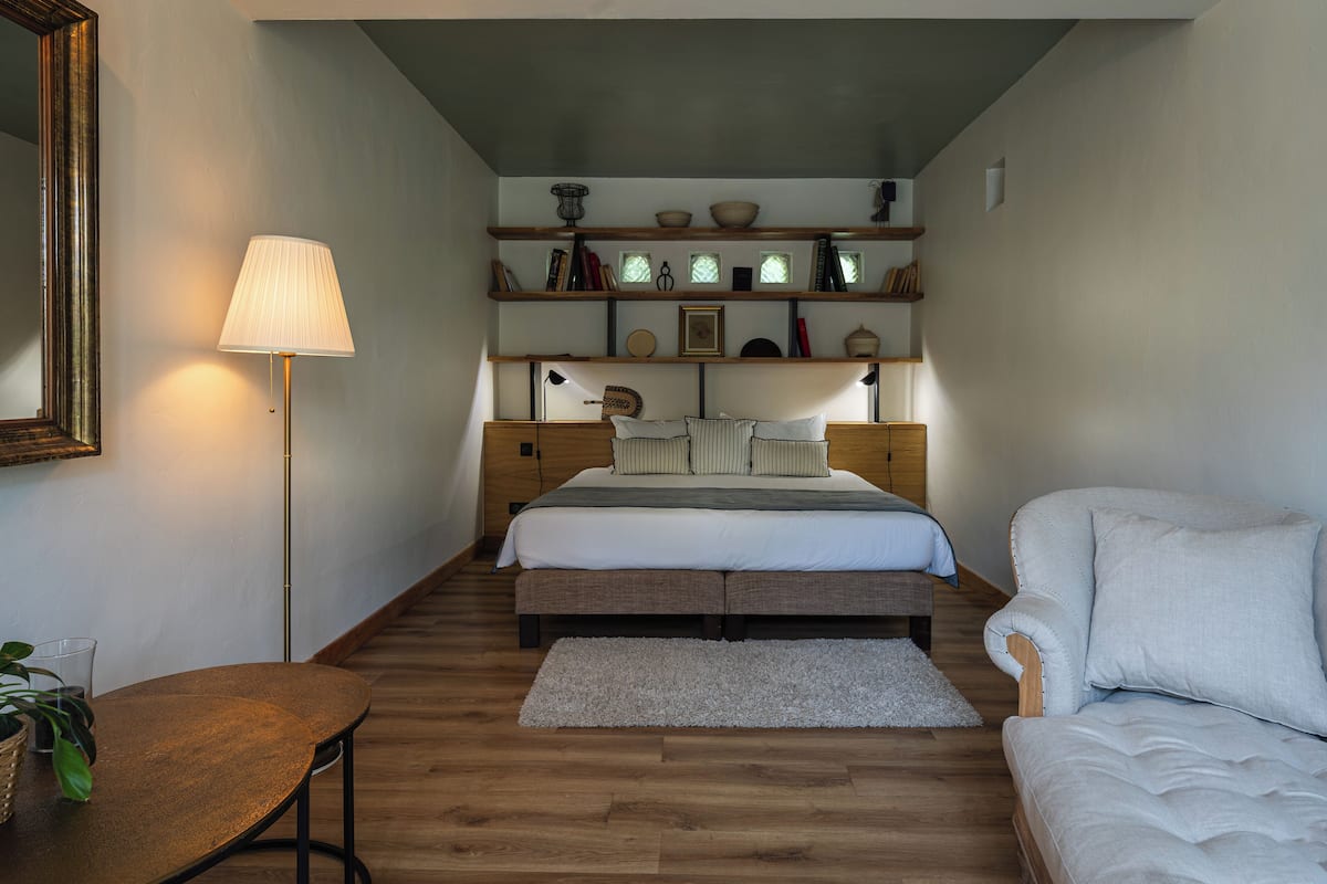 Een slaapkamer met een bed, nachtkastjes, een lamp, een sofa, een spiegel en planken met decoratieve objecten.